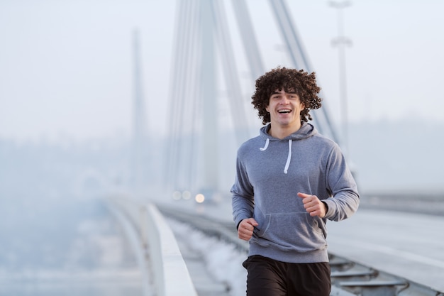 Foto il giovane uomo caucasico con capelli ricci si è vestito in abiti sportivi che corrono sul ponte all'inverno. concetto di stile di vita sano.