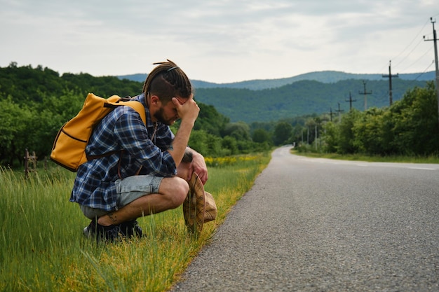 Young Caucasian man with dreadlocks and beard hitchhiking with backpack sitting on side