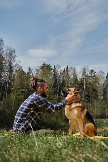 Young Caucasian man with dreadlocks is resting in park with dog Male owner strokes German Shepherd sitting on green grass in spring park and smiles Concept of pet as family member