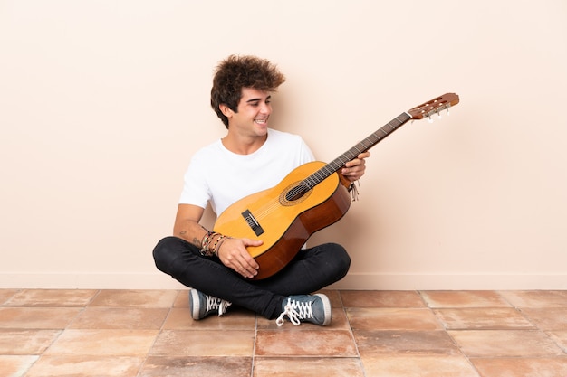 Young caucasian man with a guitar sitting on the floor with happy expression