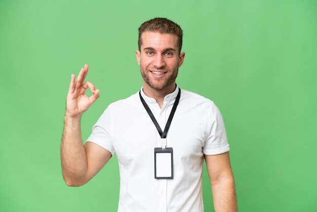 Young caucasian man with ID card isolated on green chroma background showing ok sign with fingers