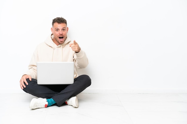 Photo young caucasian man with a laptop sitting on the floor isolated on white background surprised and pointing front