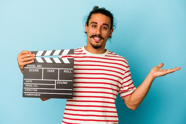 Young caucasian man with long hair holding clapperboard isolated on blue background showing a copy space on a palm and holding another hand on waist.