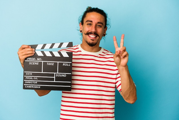 Young caucasian man with long hair holding clapperboard isolated on blue background showing number two with fingers.