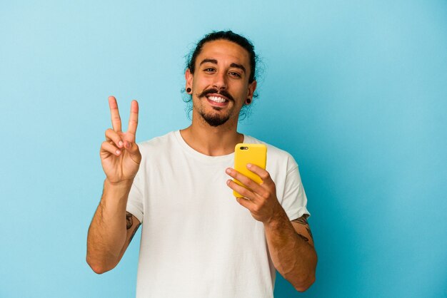 Young caucasian man with long hair holding mobile phone isolated on blue background joyful and carefree showing a peace symbol with fingers.