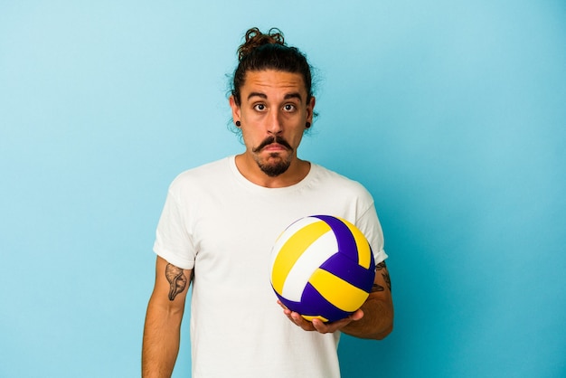 Young caucasian man with long hair isolated on blue background shrugs shoulders and open eyes confused.