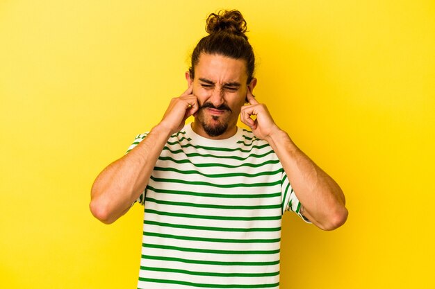 Young caucasian man with long hair isolated on yellow background covering ears with fingers, stressed and desperate by a loudly ambient.