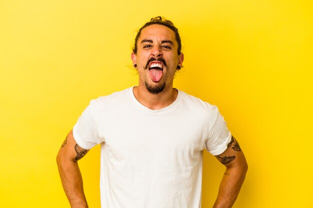 Young caucasian man with long hair isolated on yellow background funny and friendly sticking out tongue.