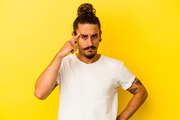 Young caucasian man with long hair isolated on yellow background pointing temple with finger, thinking, focused on a task.