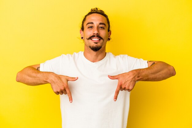 Young caucasian man with long hair isolated on yellow background points down with fingers, positive feeling.
