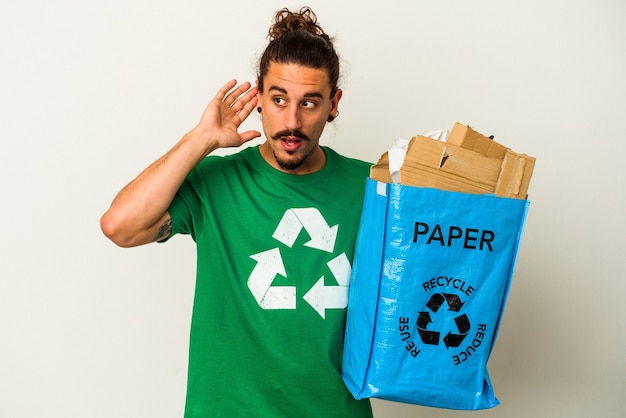 Young caucasian man with long hair recycling cardboard isolated on white background trying to listening a gossip.