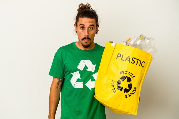 Young caucasian man with long hair recycling plastic isolated on white background shrugs shoulders and open eyes confused.