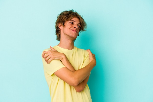 Giovane uomo caucasico con trucco isolato su sfondo blu abbracci, sorridendo spensierato e felice.