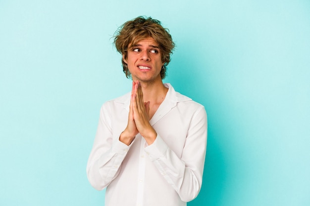 Young caucasian man with make up isolated on blue background  praying, showing devotion, religious person looking for divine inspiration.