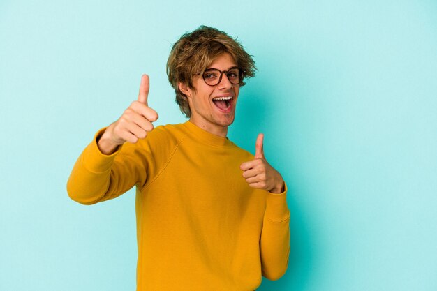 Young caucasian man with make up isolated on blue background  raising both thumbs up, smiling and confident.