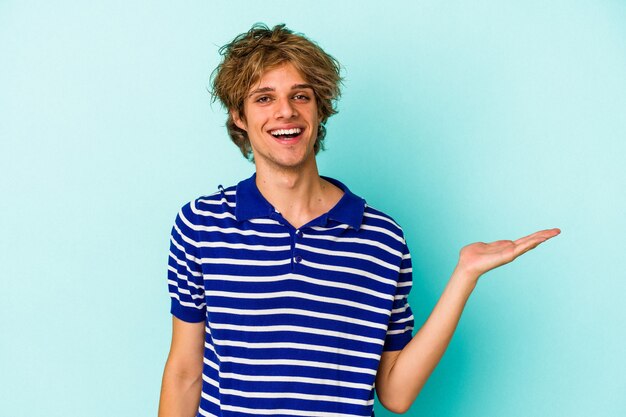 Young caucasian man with make up isolated on blue background  showing a copy space on a palm and holding another hand on waist.