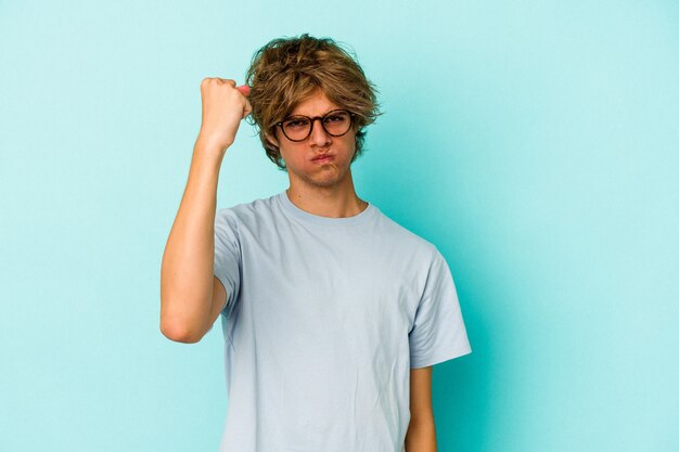 Young caucasian man with make up isolated on blue background  showing fist to camera, aggressive facial expression.