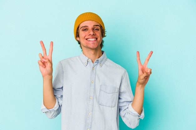 Young caucasian man with make up isolated on blue background  showing victory sign and smiling broadly.