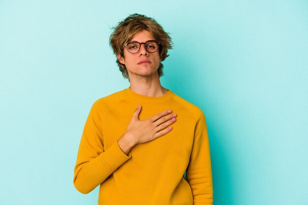 Young caucasian man with make up isolated on blue background  taking an oath, putting hand on chest.