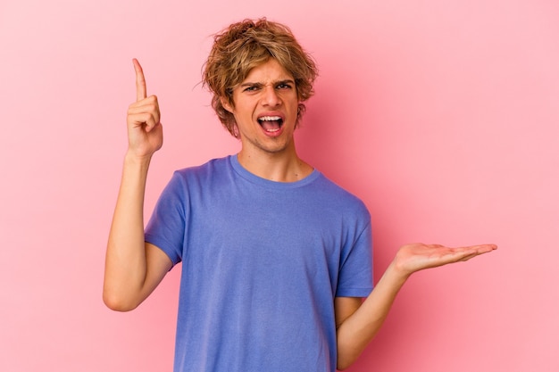 Young caucasian man with make up isolated on pink background holding and showing a product on hand.