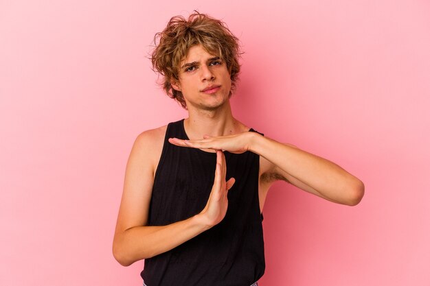 Young caucasian man with make up isolated on pink background showing a timeout gesture.