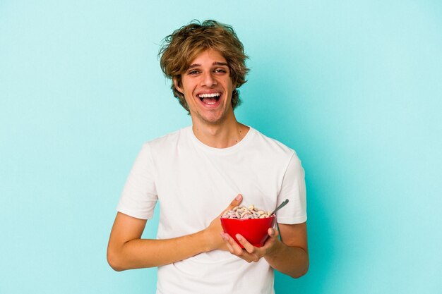 Young caucasian man with makeup a cereal bowl isolated on blue background  laughing and having fun.