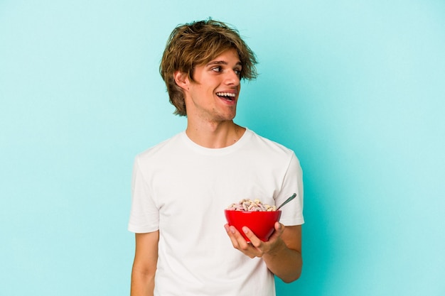 Young caucasian man with makeup a cereal bowl isolated on blue background  looks aside smiling, cheerful and pleasant.
