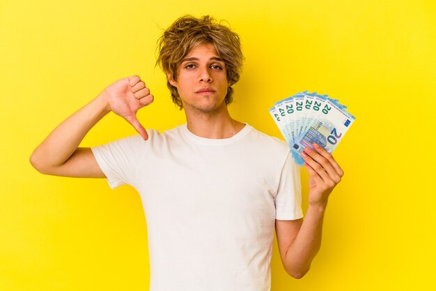 Young caucasian man with makeup holding bills isolated on yellow background  showing a dislike gesture, thumbs down. Disagreement concept.