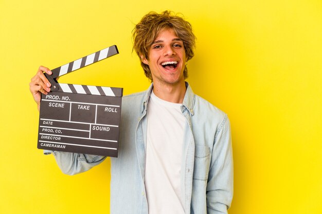 Young caucasian man with makeup holding clapperboard isolated on yellow background  happy, smiling and cheerful.