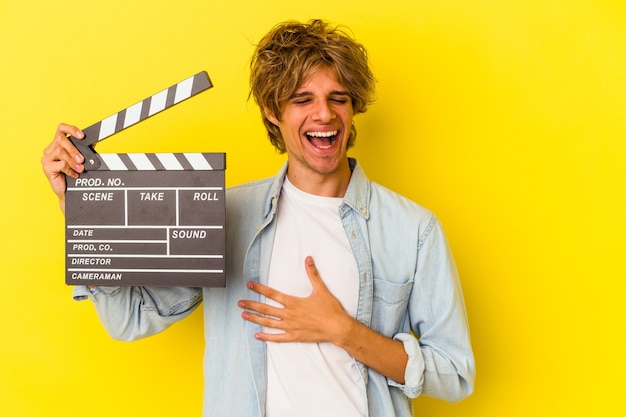 Young caucasian man with makeup holding clapperboard isolated on yellow background  laughs out loudly keeping hand on chest.