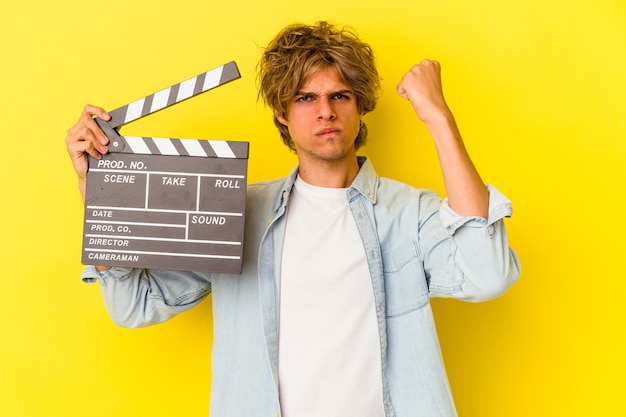 Giovane uomo caucasico con il ciak della tenuta di trucco isolato su fondo giallo che mostra il pugno alla macchina fotografica, espressione facciale aggressiva.