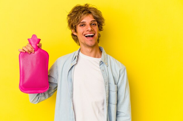young caucasian man with makeup holding a hot bag of water isolated on yellow background  happy, smiling and cheerful.