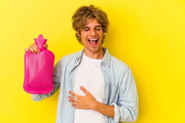 Young caucasian man with makeup holding a hot bag of water isolated on yellow background  laughing and having fun.