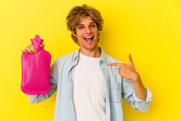 Young caucasian man with makeup holding a hot bag of water isolated on yellow background  person pointing by hand to a shirt copy space, proud and confident