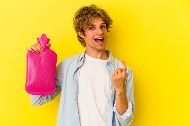 young caucasian man with makeup holding a hot bag of water isolated on yellow background  pointing with finger at you as if inviting come closer.