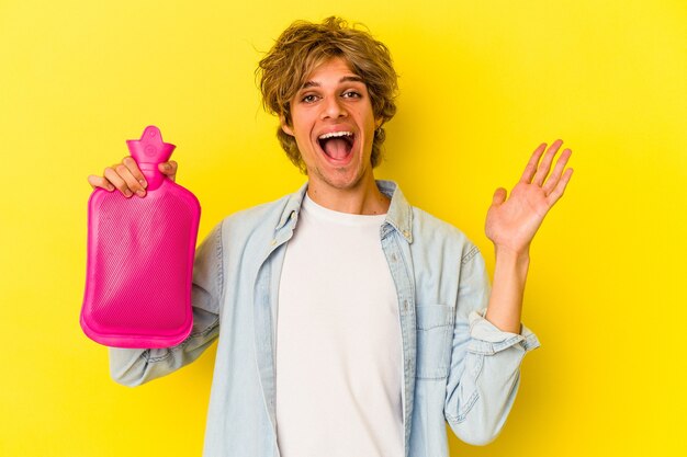 Young caucasian man with makeup holding a hot bag of water isolated on yellow background  receiving a pleasant surprise, excited and raising hands.