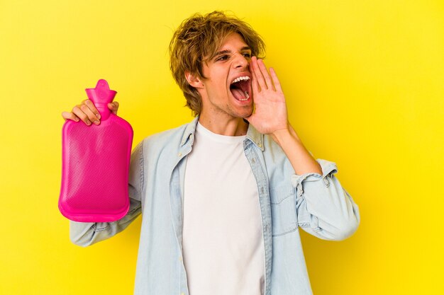 Young caucasian man with makeup holding a hot bag of water isolated on yellow background  shouting and holding palm near opened mouth.