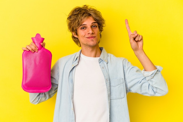 Giovane uomo caucasico con il trucco che tiene una borsa d'acqua calda isolata su sfondo giallo che mostra il numero uno con il dito.