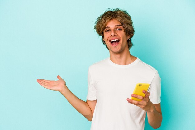 Young caucasian man with makeup holding mobile phone isolated on blue background  showing a copy space on a palm and holding another hand on waist.