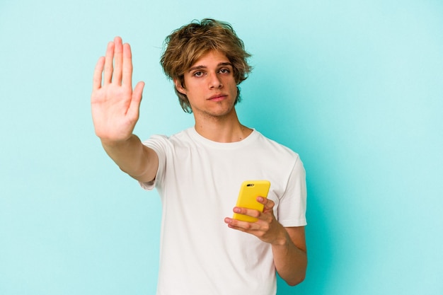 Young caucasian man with makeup holding mobile phone isolated on blue background  standing with outstretched hand showing stop sign, preventing you.