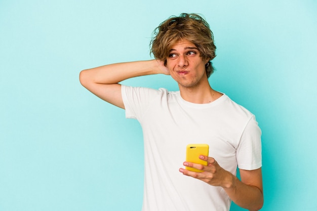 Giovane uomo caucasico con il trucco che tiene il telefono cellulare isolato su sfondo blu toccando la parte posteriore della testa, pensando e facendo una scelta.