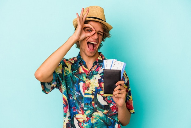 Young caucasian man with makeup holding passport isolated on blue background  excited keeping ok gesture on eye.