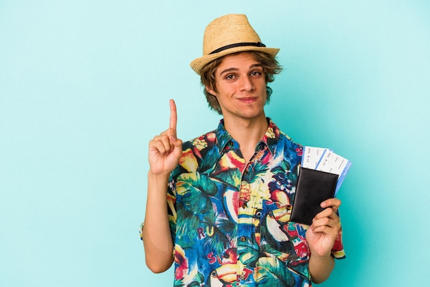 Young caucasian man with makeup holding passport isolated on blue background  showing number one with finger.