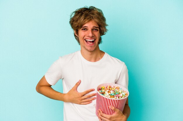 Young caucasian man with makeup holding popcorn isolated on blue background  laughing and having fun.