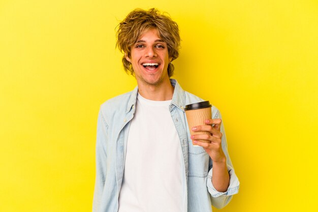 Young caucasian man with makeup holding a take away coffee isolated on yellow background  happy, smiling and cheerful.