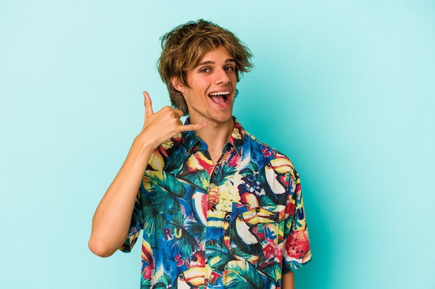 Young caucasian man with makeup wearing a summer clothes isolated on blue background  showing a mobile phone call gesture with fingers.