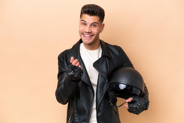 お金のジェスチャーを作るベージュ色の背景に分離されたオートバイのヘルメットを持つ若い白人男