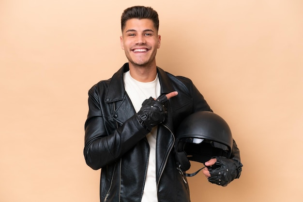 製品を提示する側を指しているベージュの背景に分離されたオートバイのヘルメットを持つ若い白人男性