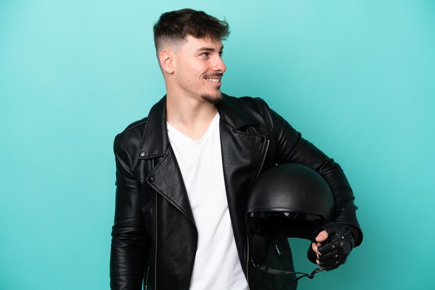 Young caucasian man with a motorcycle helmet isolated on blue background looking to the side and smiling