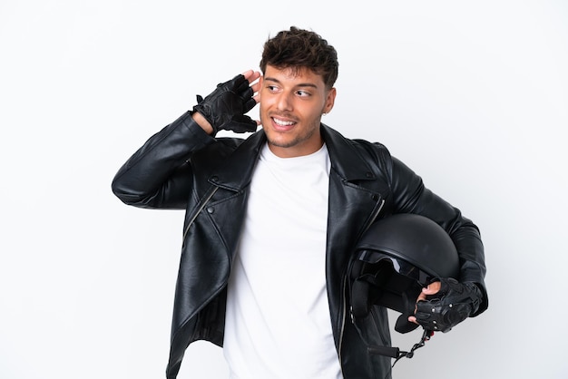 Young caucasian man with a motorcycle helmet isolated on white background listening to something by putting hand on the ear